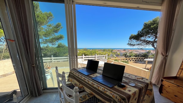Een kamer met twee laptops op een tafel, een stoel en uitzicht op zee en stad door grote ramen.