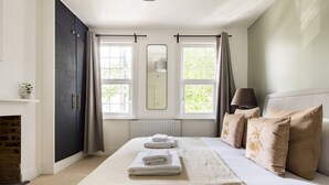 Un dormitorio con una cama, dos ventanas, un espejo, un radiador y una lámpara.