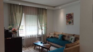 Un salon avec un canapé, une table basse et un téléviseur. Il y a une fenêtre avec des rideaux et un tableau accroché au mur.