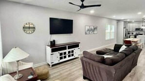 A modern living room with a sofa, a TV, a ceiling fan, and a kitchen area in the background.