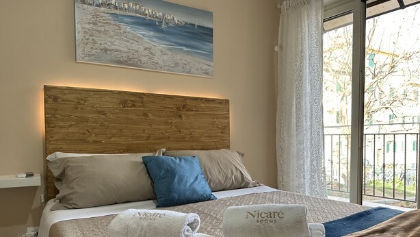 Une chambre d’hôtel avec une tête de lit en bois, un tableau représentant un paysage côtier, un balcon avec vue et deux serviettes portant le nom de l’hôtel.