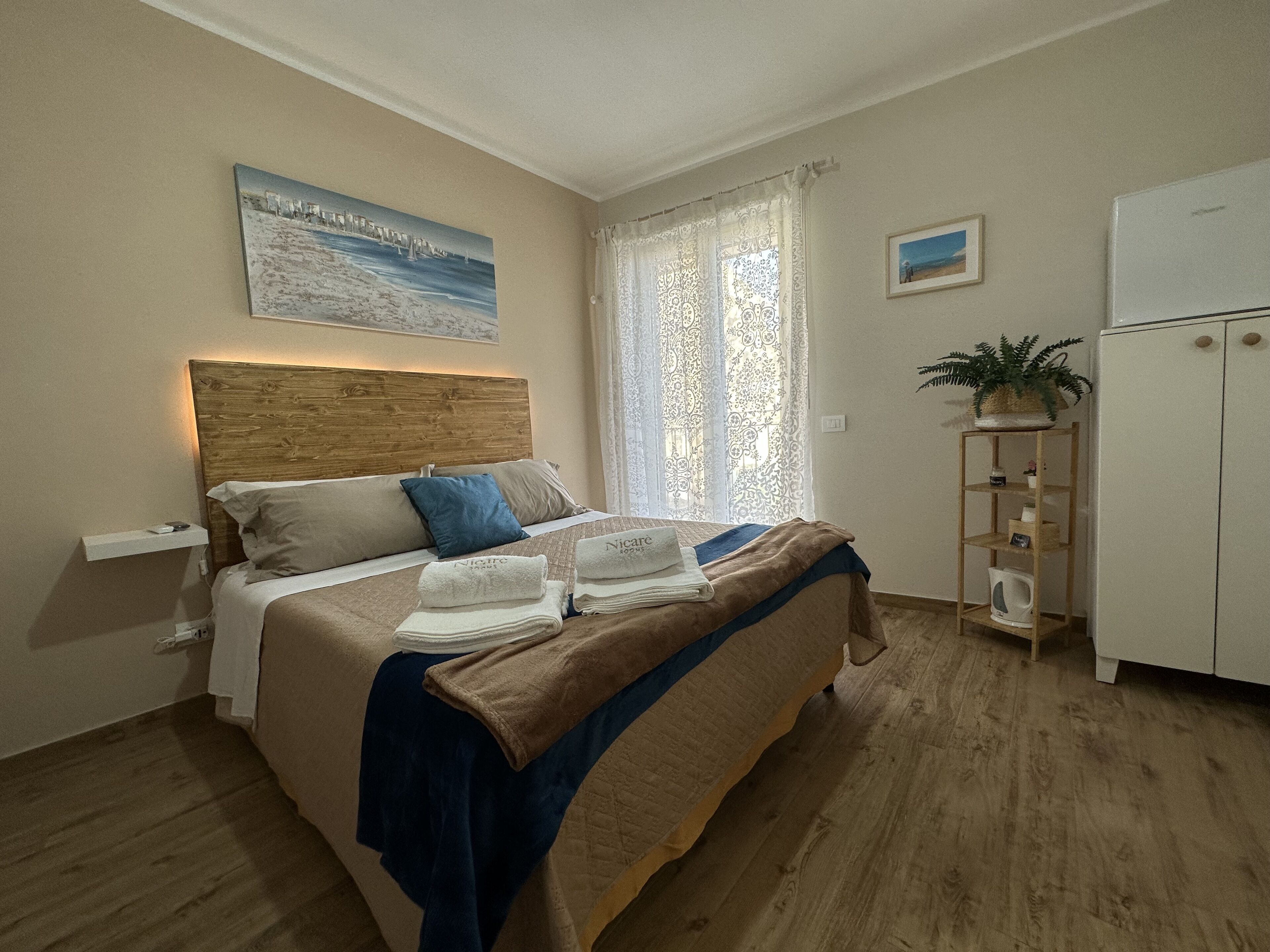 A bedroom with a wooden headboard, a bed with beige and blue bedding, a nightstand, a window with patterned curtains, a small shelf with a plant, and a white wardrobe.