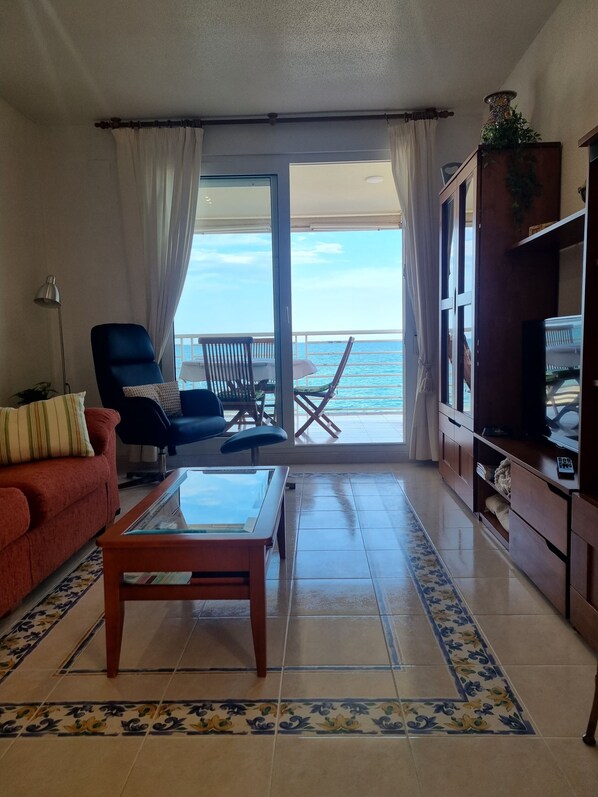 En stue med sofa, lænestol og sofabord. Der er en balkon med udsigt over havet.