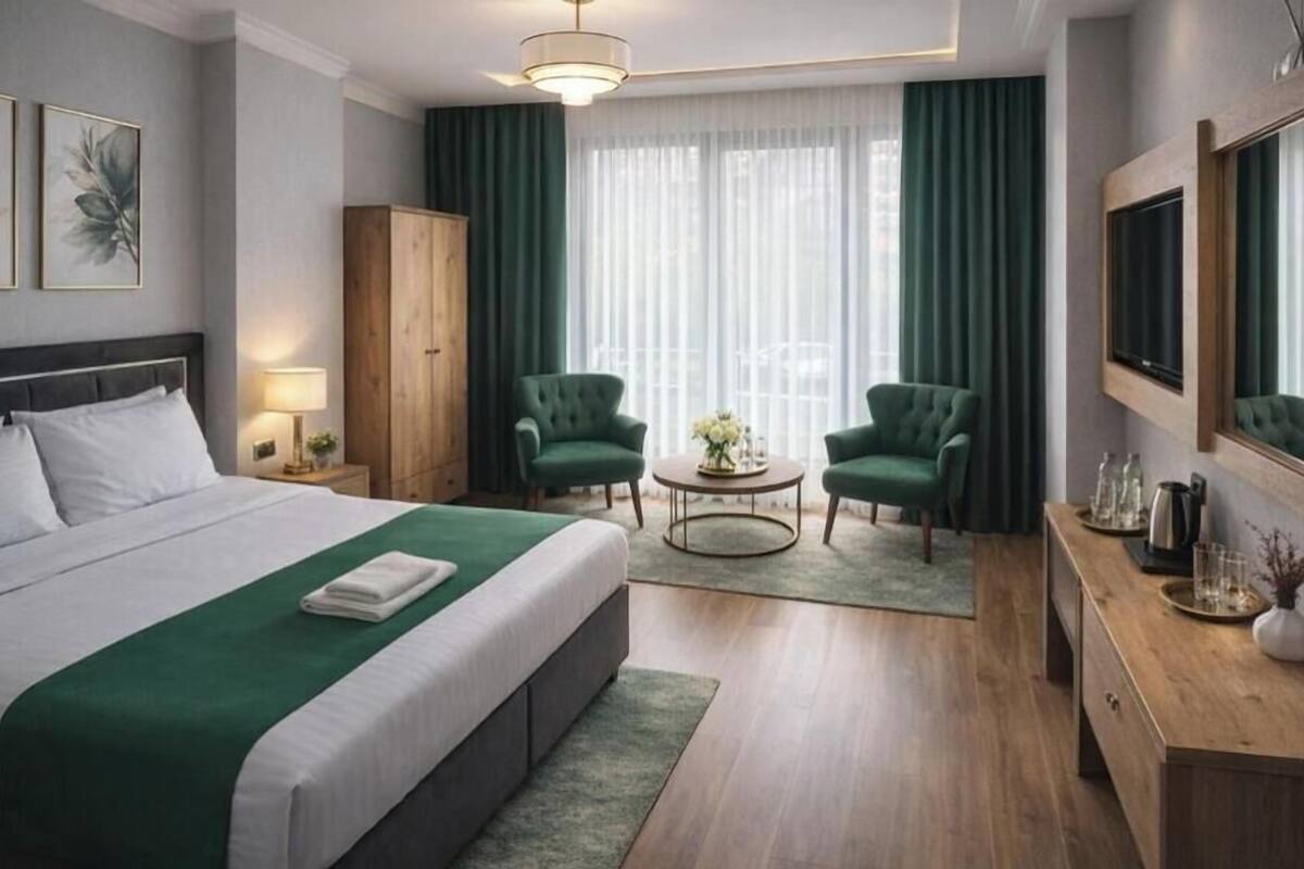 Ein Hotelzimmer mit einem Bett, zwei grünen Sessel, einem runden Couchtisch, einem Holzschrank, einem Fernseher und einem großen Fenster mit Vorhängen.