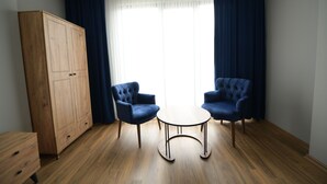Une pièce avec une armoire en bois, deux fauteuils bleus, une petite table ronde et une grande fenêtre avec des rideaux.