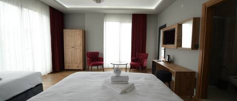 A hotel room with a large bed, a wooden wardrobe, two red armchairs, a small round table, and a TV mounted on the wall.