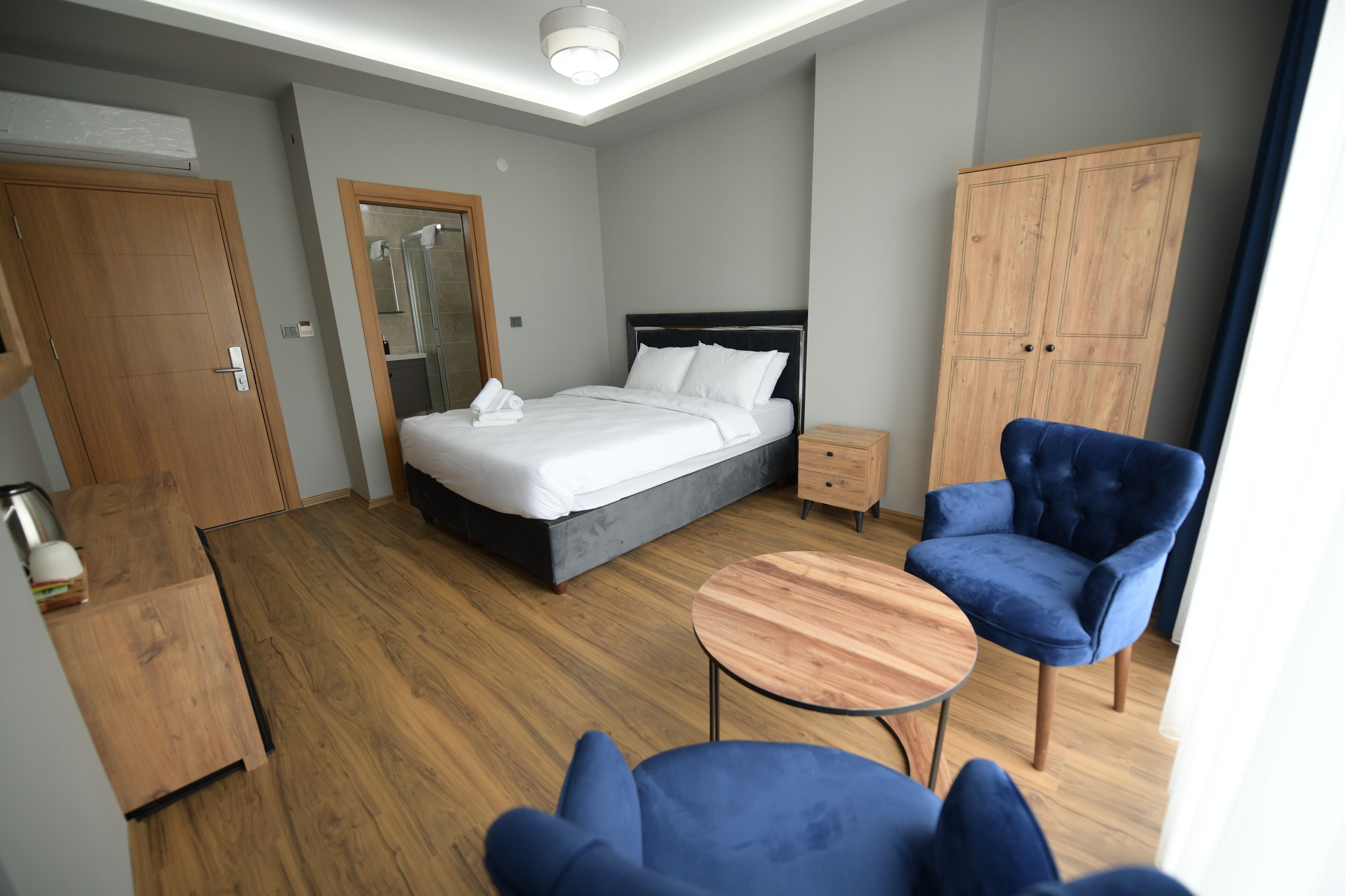 A hotel room with a bed, a wooden wardrobe, a blue armchair, a small wooden table, and a shower area.