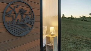 A room with a wooden wall, a circular metal artwork, a bedside table with a lamp, and a view of a grassy field. - The High Country Views Escape by Tiny Away (Table Top)