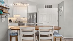 A modern kitchen with white cabinets, a stainless steel refrigerator, and a dining area with a wooden table and chairs. - Updated 2BR/2BA Condo in North Ocean City, MD | Indoor + Outdoor Pools | 2 Blocks to Beach | Walk to Food Lion & Restaurants | Sleeps 6 (Ocean City)