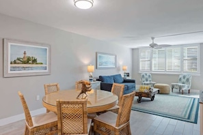 A dining area with wicker chairs and a glass table, a living room with a sofa and patterned chairs, and a view of the ocean through large windows.