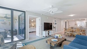 A spacious living area with a large sliding glass door, a ceiling fan, a television, a dining table with chairs, and a blue sofa.