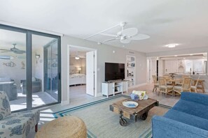 A spacious living area with a large sliding glass door, a ceiling fan, a television, a dining table with chairs, and a blue sofa.