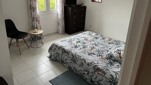 Une chambre à coucher avec un lit, une chaise, une petite table, une commode et une fenêtre avec des rideaux.