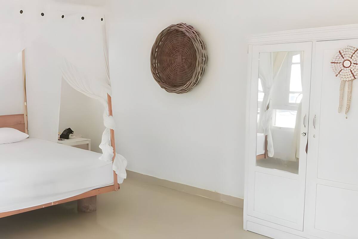 Ein weißes Himmelbett mit Holz-Kopfstück, ein Nachttisch mit Lampe und ein Kleiderschrank mit Spiegel.