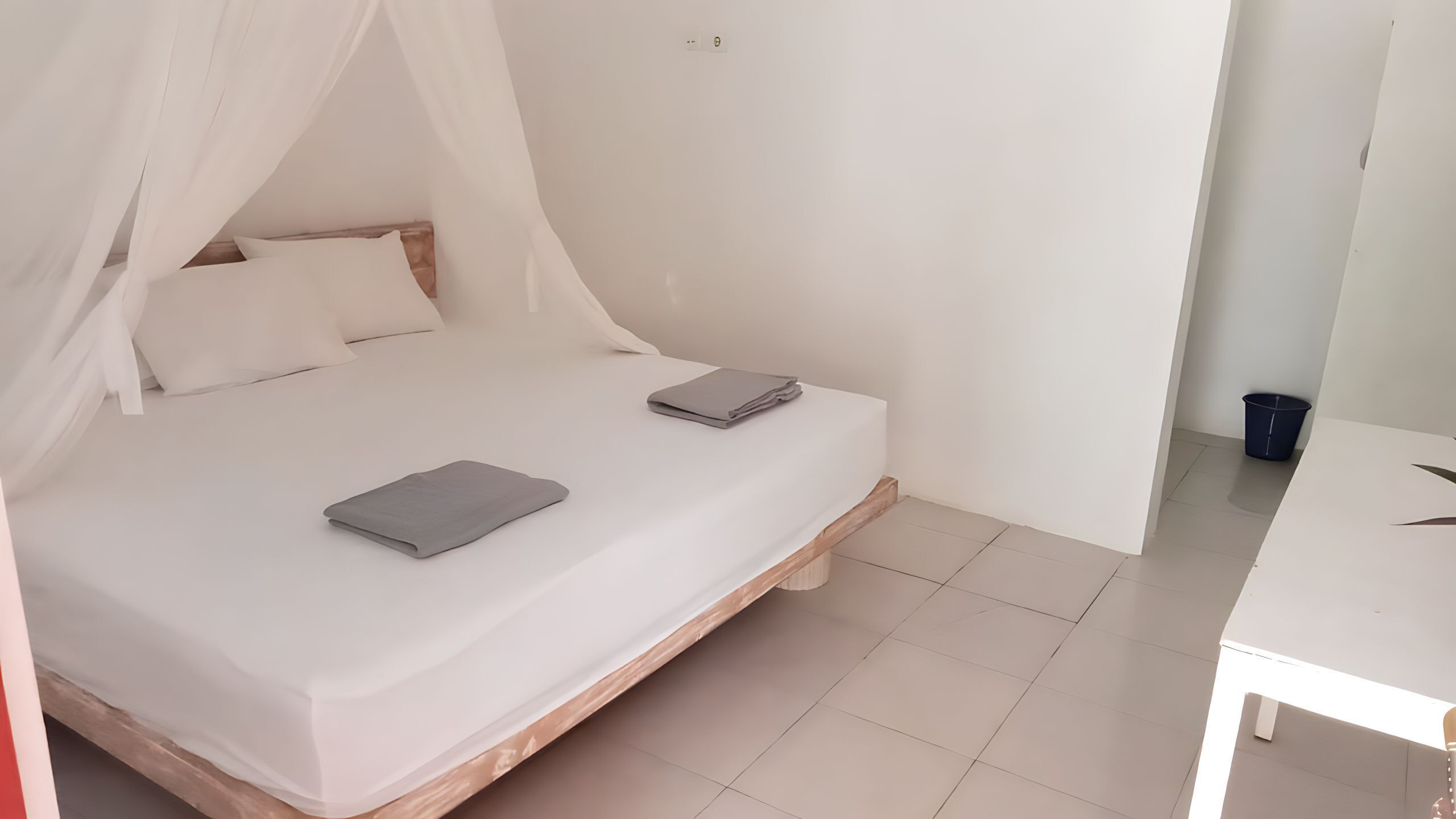 Un lit simple surmonté d’un baldaquin blanc, une petite table de chevet et une poubelle bleue dans une pièce au décor minimaliste.