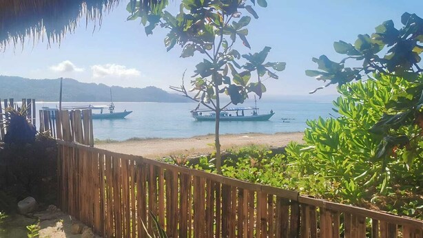 A wooden fence, a thatched roof, and a view of boats on a calm sea with distant mountains. - THE VILLAGE INN Rua Beach  (Wanokaka)