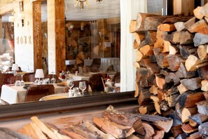 A restaurant with wooden decor, tables set with glasses and cutlery, and a stack of firewood in the foreground.