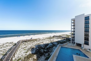 A beach view from a hotel with a pool, a wooden boardwalk, and a white building with balconies.