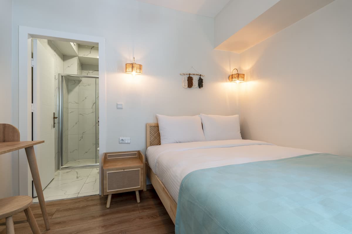 A bedroom with a bed, bedside tables, a nightstand, a wooden chair, a glass shower enclosure, and wall-mounted lights.