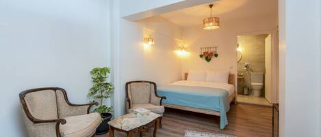 Quarto com cama, duas poltronas, uma mesinha, uma planta em vaso e um tapete estampado.