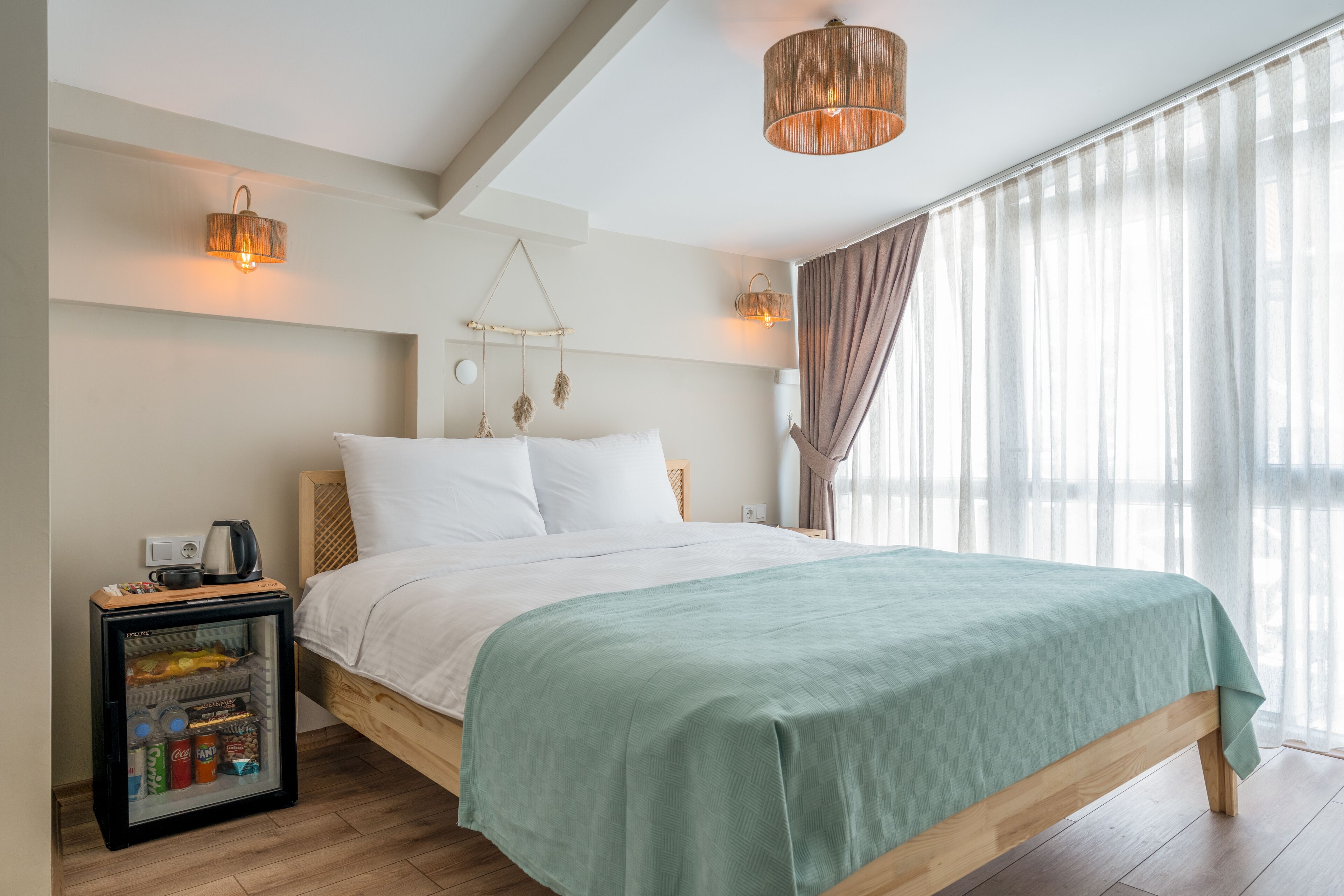 Un dormitorio con cama, mesita de noche con lámpara, refrigerador pequeño y cortinas translúcidas.