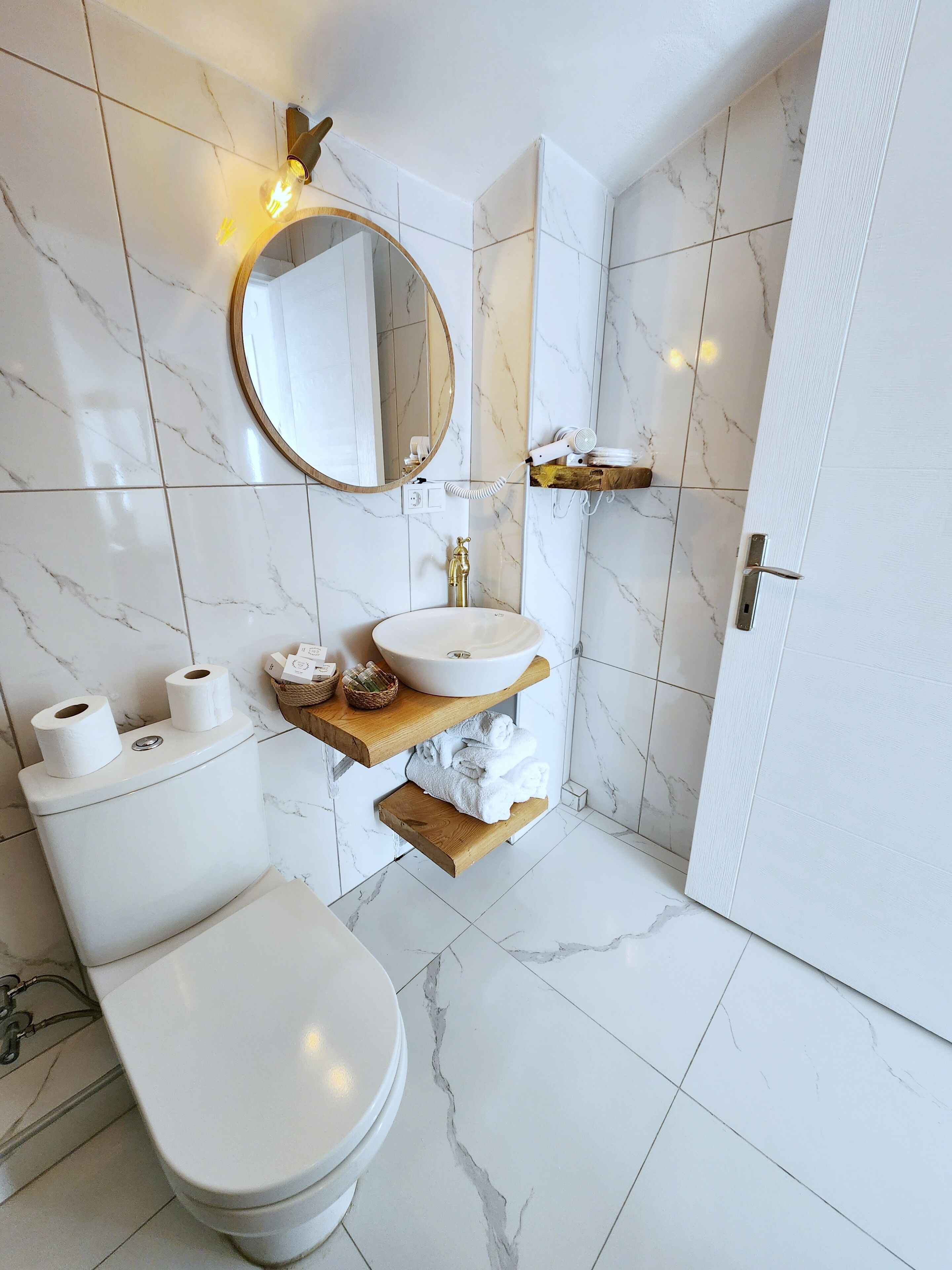 Une salle de bain moderne avec des toilettes blanches, un miroir rond, un lavabo avec un plan de toilette en marbre et une étagère en bois où sont posées des serviettes.