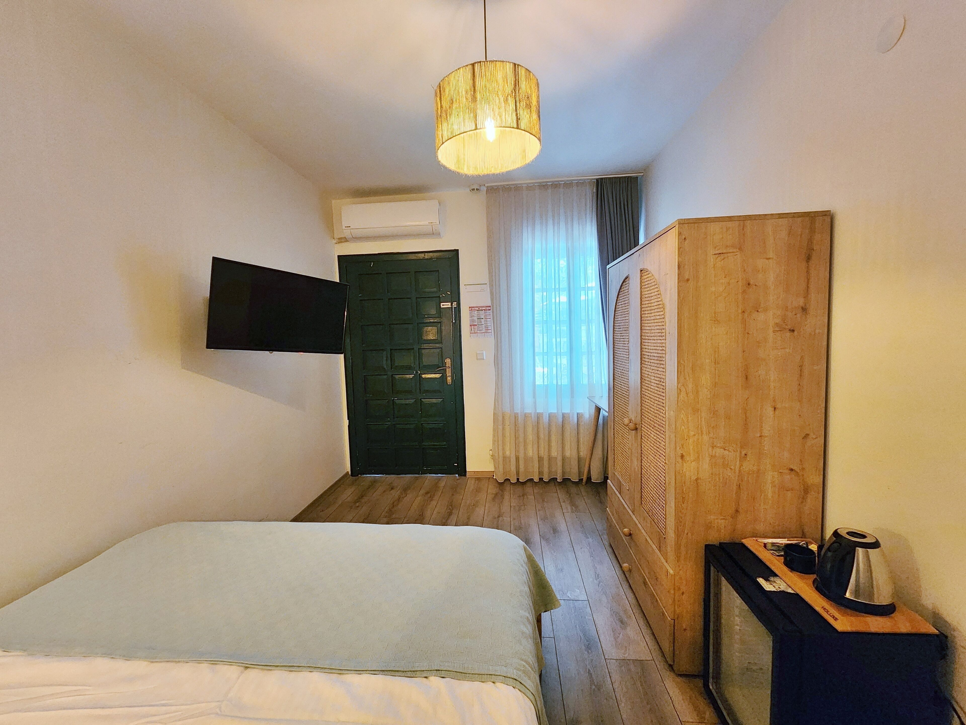 Ett hotellrum med en säng, en tv som är monterad på väggen, en garderob i trä och ett skrivbord med en vattenkokare.