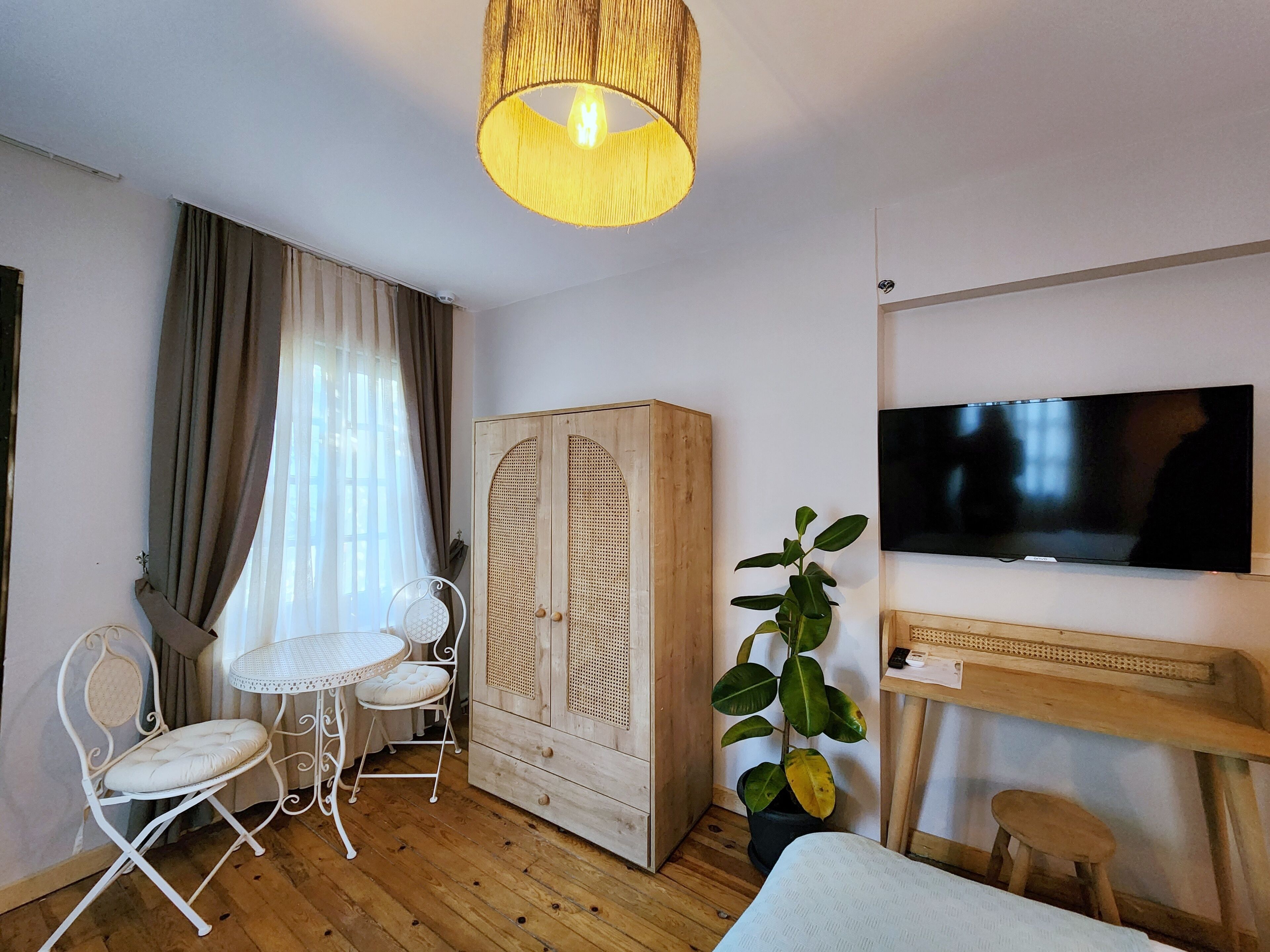 A room with a wooden wardrobe, a TV mounted on the wall, a small table with chairs, and a potted plant.