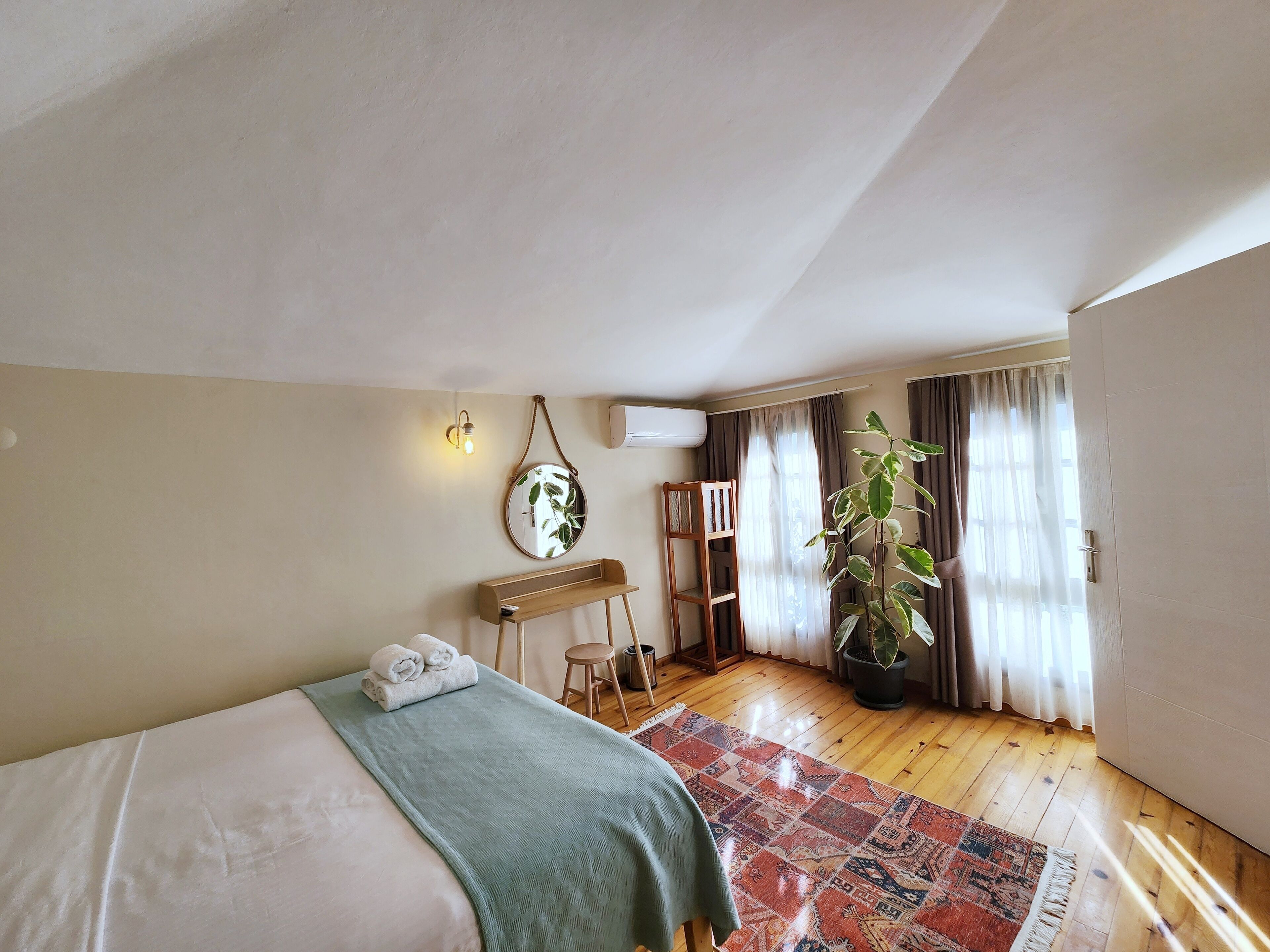 A bedroom with a bed, a small table, a chair, a potted plant, and a window with curtains.
