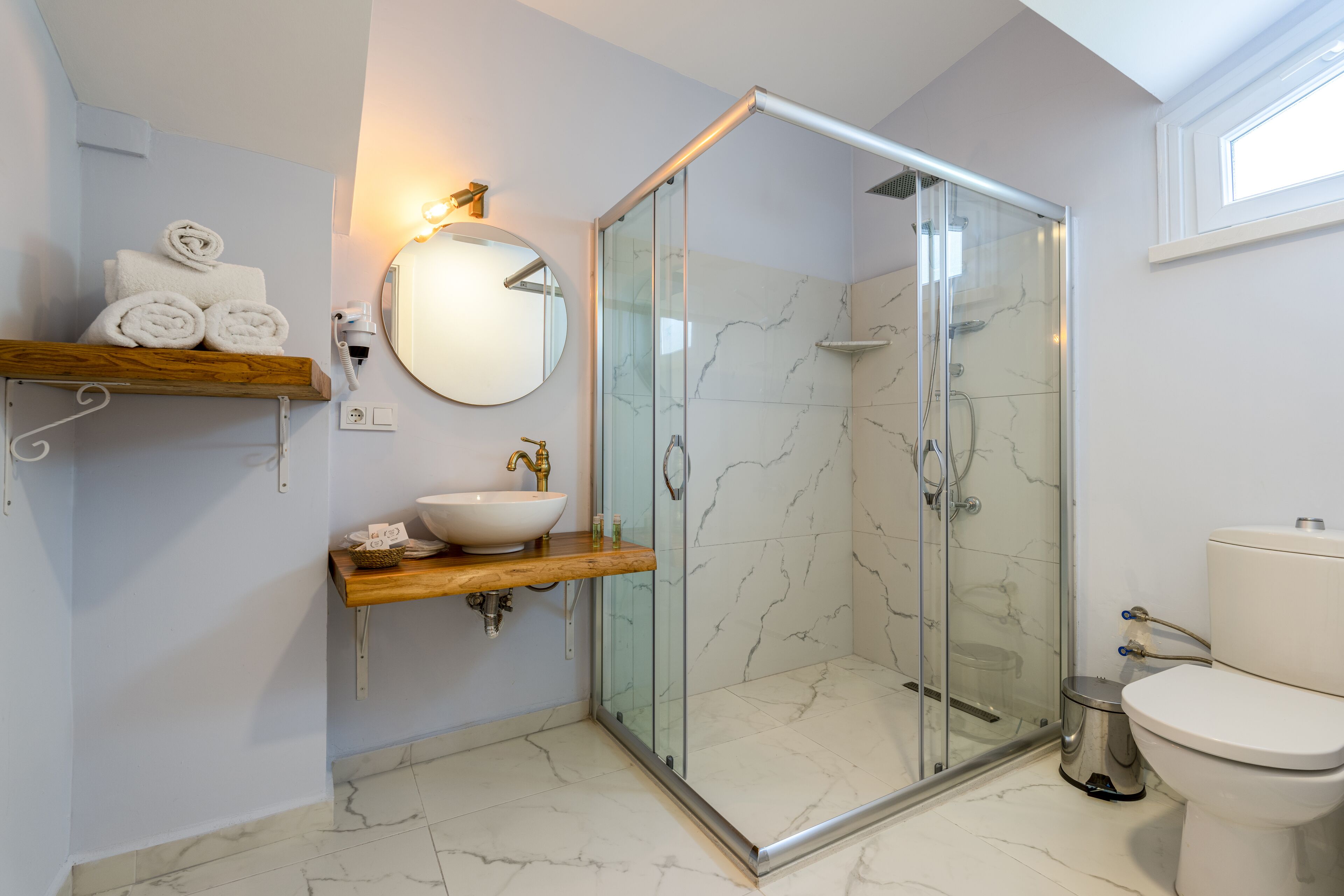 A modern bathroom with a glass-enclosed shower, a wooden shelf with towels, a round mirror, and a white toilet.