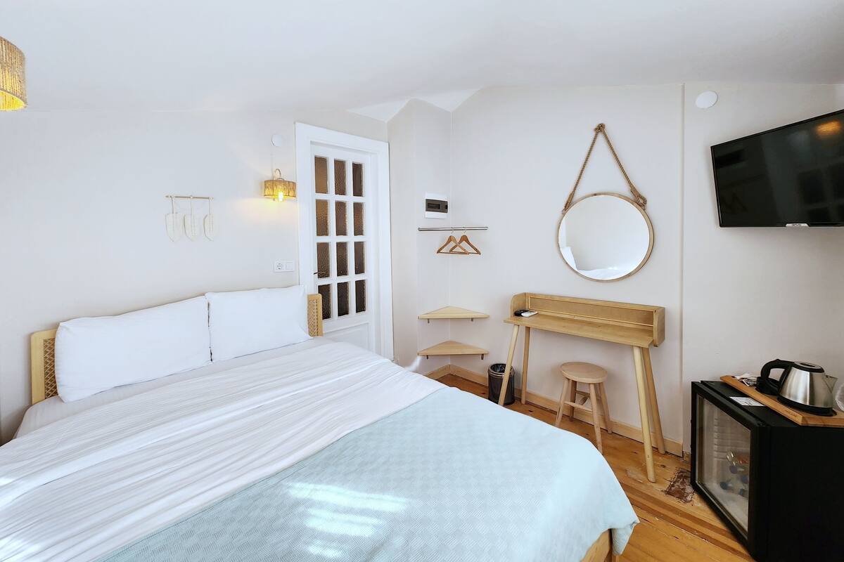 A bedroom with a bed, a desk, a mirror, a TV, and a small stool.