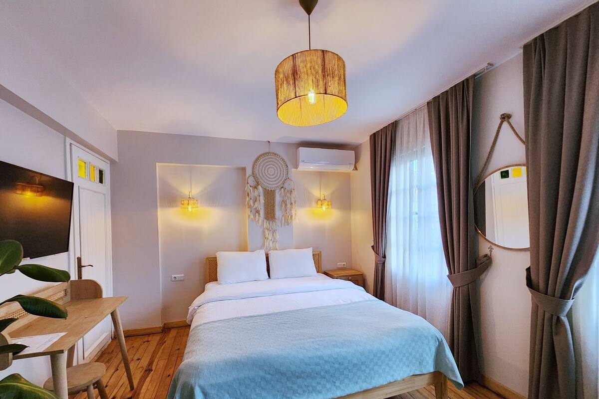A bedroom with a bed, bedside lamps, a wall-mounted air conditioner, a mirror, and a wooden desk with a chair.