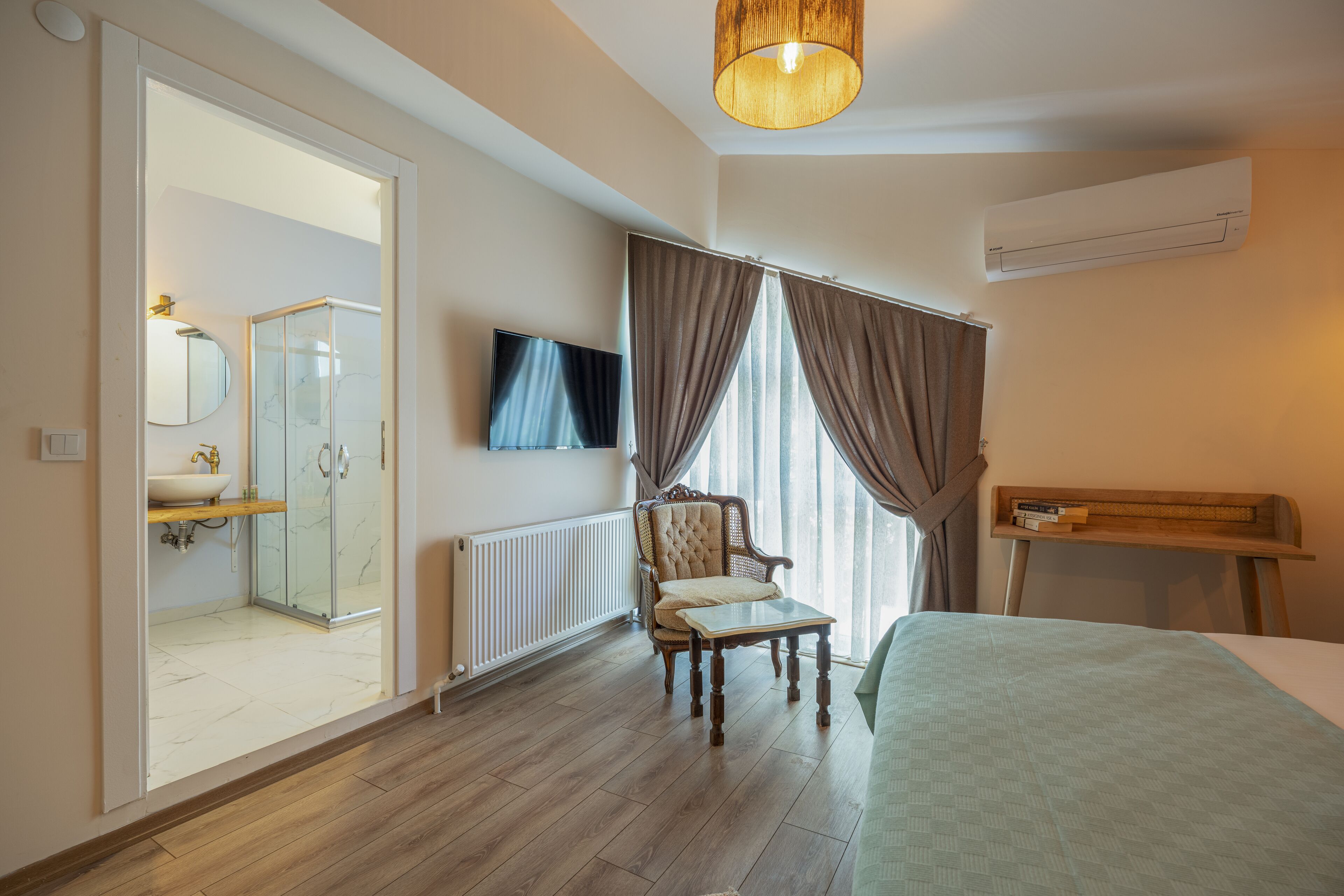 A bedroom with a bed, a chair, a TV, a window with curtains, and a bathroom with a shower and a mirror.