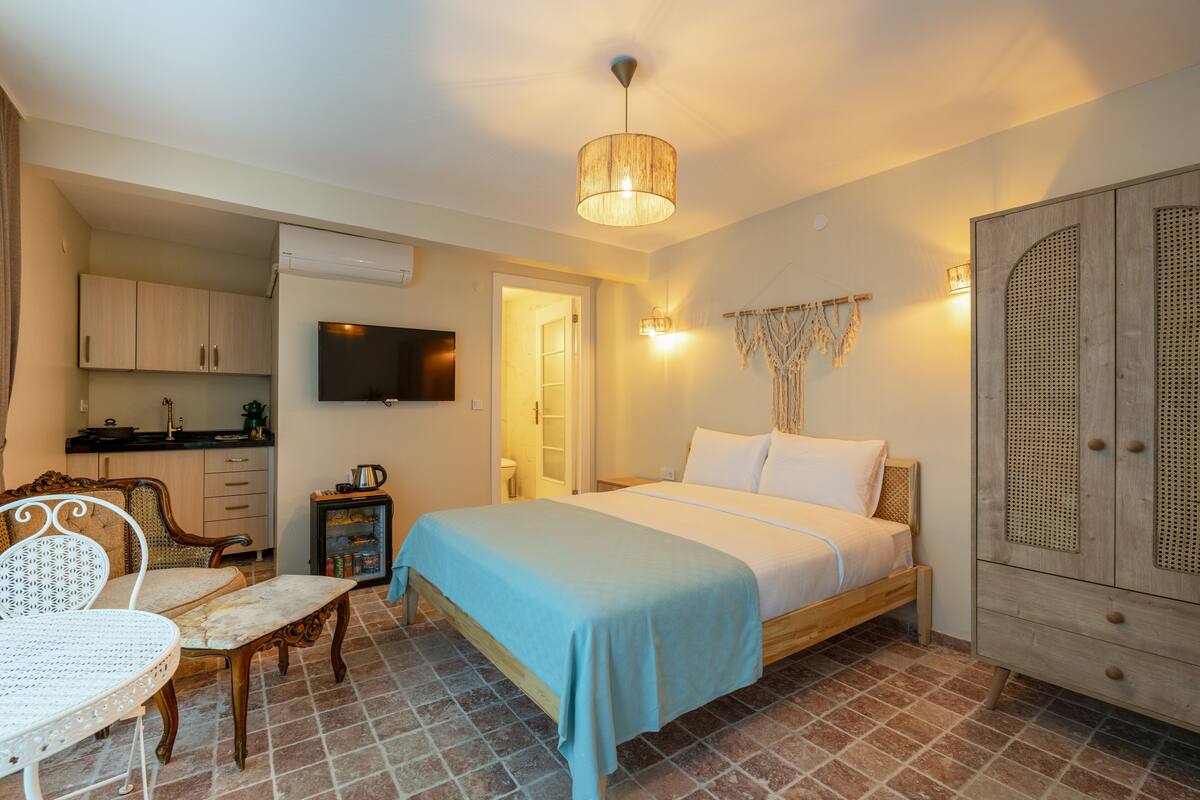 A bedroom with a bed, a chair, a table, a wardrobe, a television, and a kitchen area.