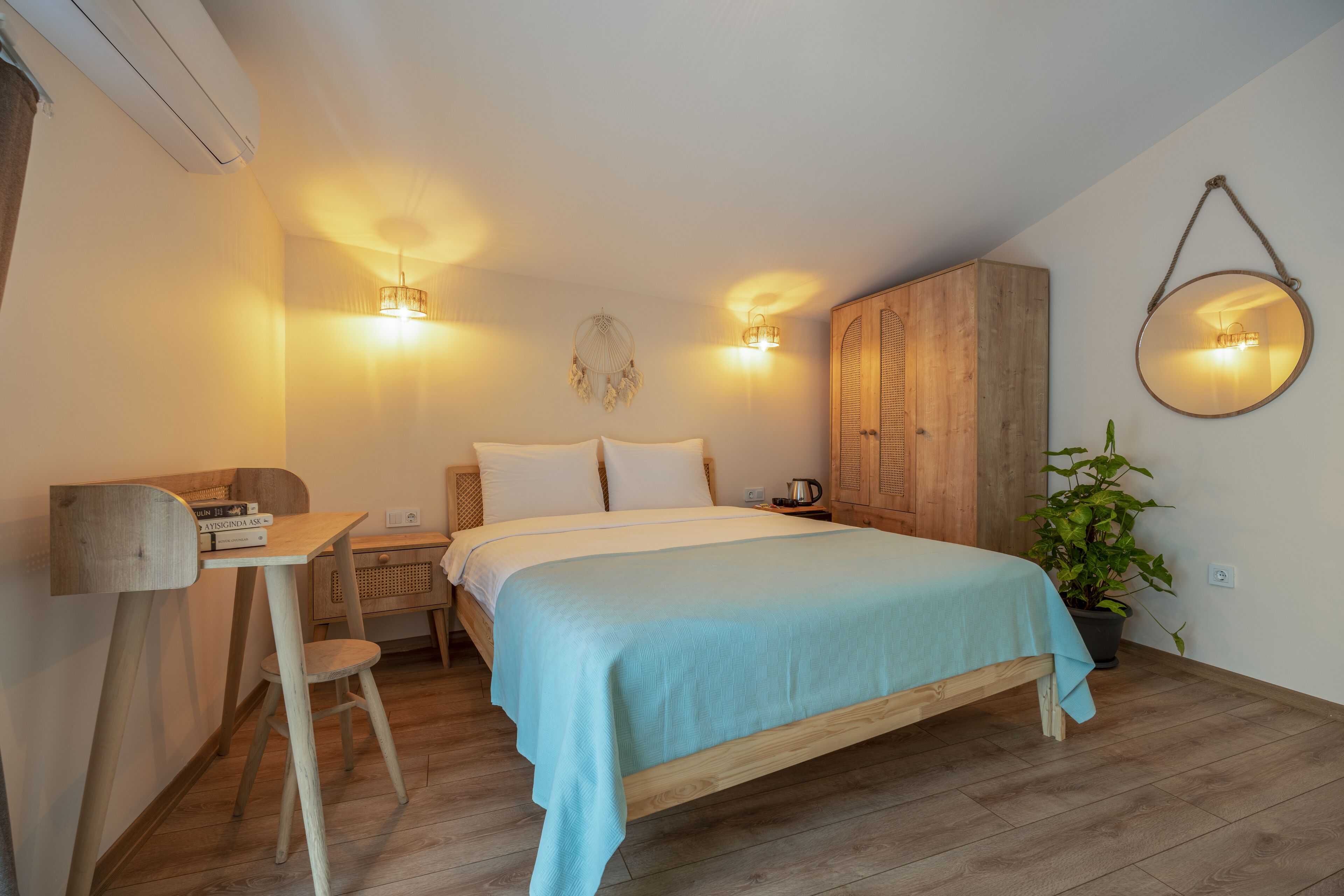 Un dormitorio con una cama de madera, un escritorio con libros, una silla, una planta en maceta y un espejo colgado.