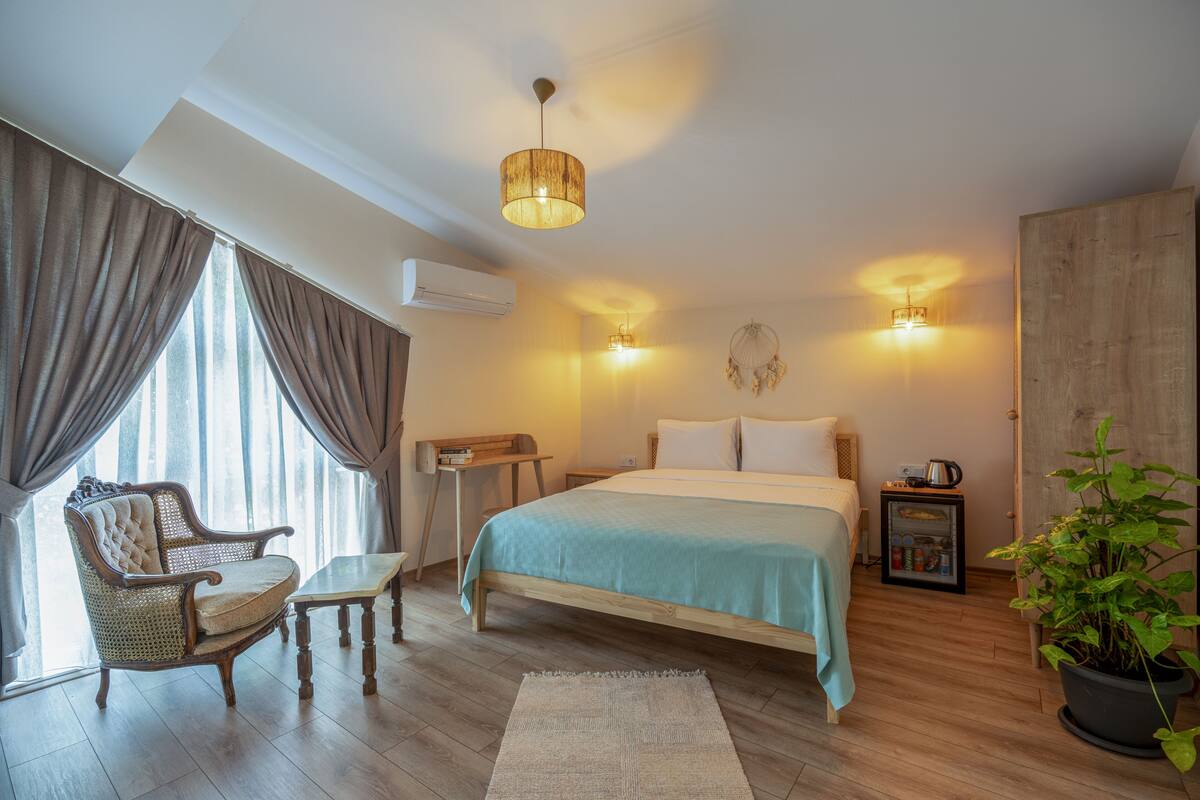 A bedroom with a bed, a chair, a bedside table, a nightstand, a lamp, a plant, and a window with curtains.