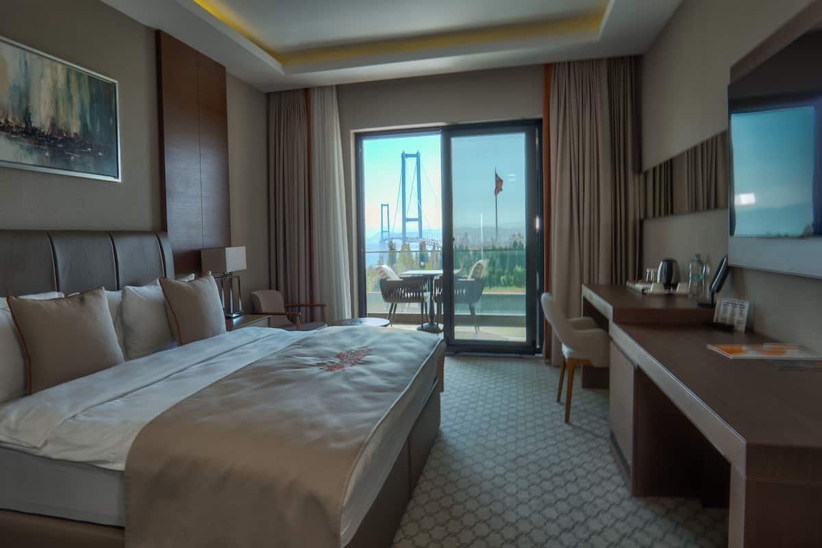 A hotel room with a large bed, a desk, a chair, a TV, and a balcony with a view of a bridge.