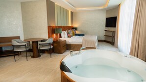 A hotel room with a large bathtub, a dining table with chairs, a bed with a TV, and a TV on the wall. - Motel Heksapolis (Dilovasi)