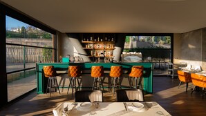 A modern bar area with a green bar counter, leather bar stools, and a variety of bottles. There are dining tables with chairs and a view of a cityscape through large windows. - Motel Heksapolis (Dilovasi)