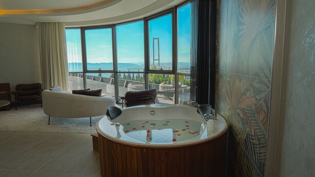 A modern bathroom with a freestanding bathtub, a large window offering a view of a bridge and the sea, and a seating area with a sofa and chairs. - Motel Heksapolis (Dilovasi)