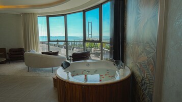 A modern bathroom with a freestanding bathtub, a large window offering a view of a bridge and the sea, and a seating area with a sofa and chairs.