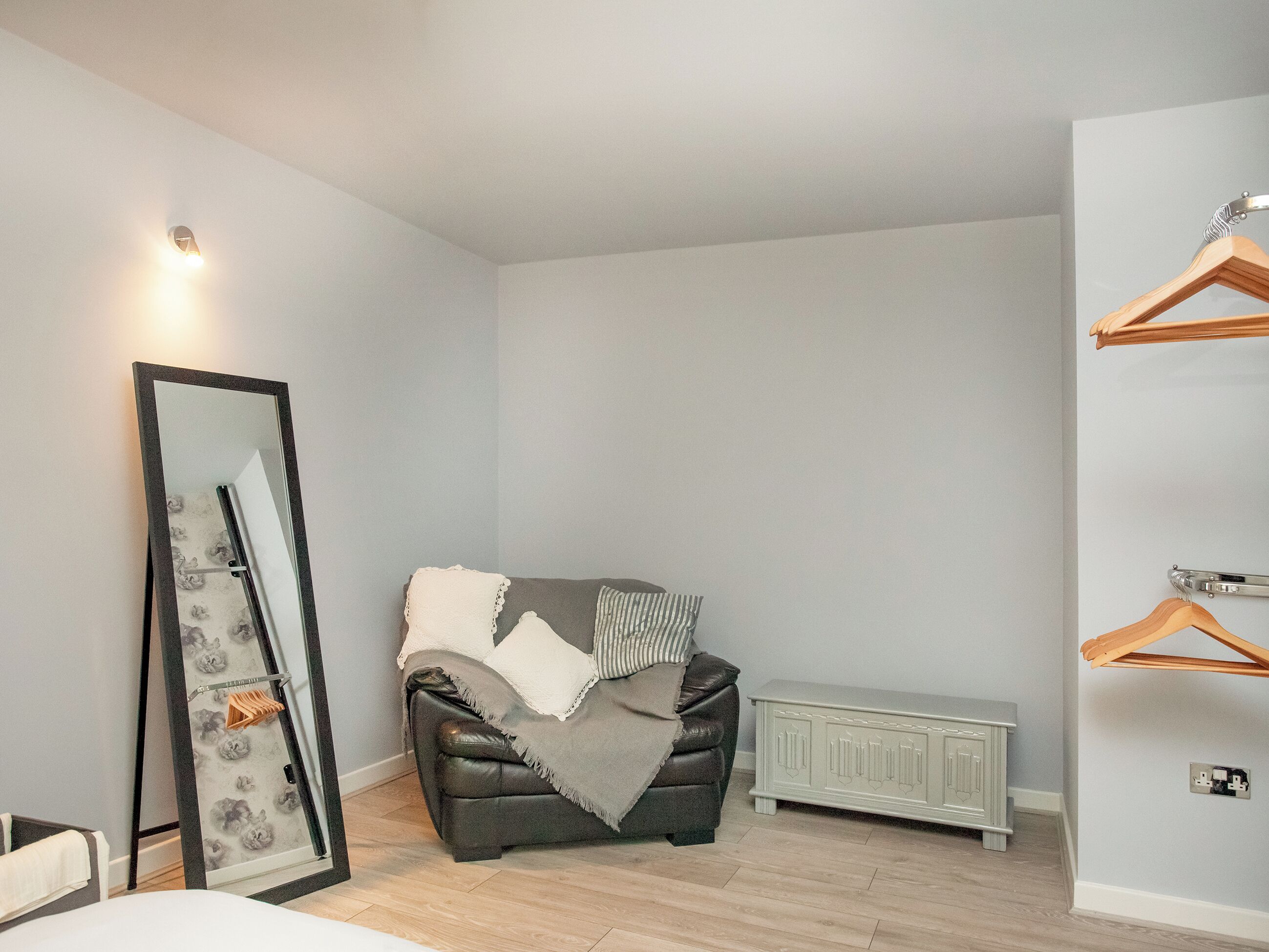 Ein modernes Schlafzimmer mit grauem Sofa, einem großen Spiegel, einem Heizkörper und Holzaufhänger an der Wand.