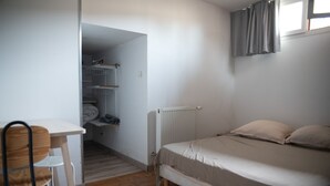 Une petite chambre avec un lit simple, un plancher en bois, un bureau blanc et une chaise. Il y a un radiateur sous le lit et une fenêtre avec des rideaux.