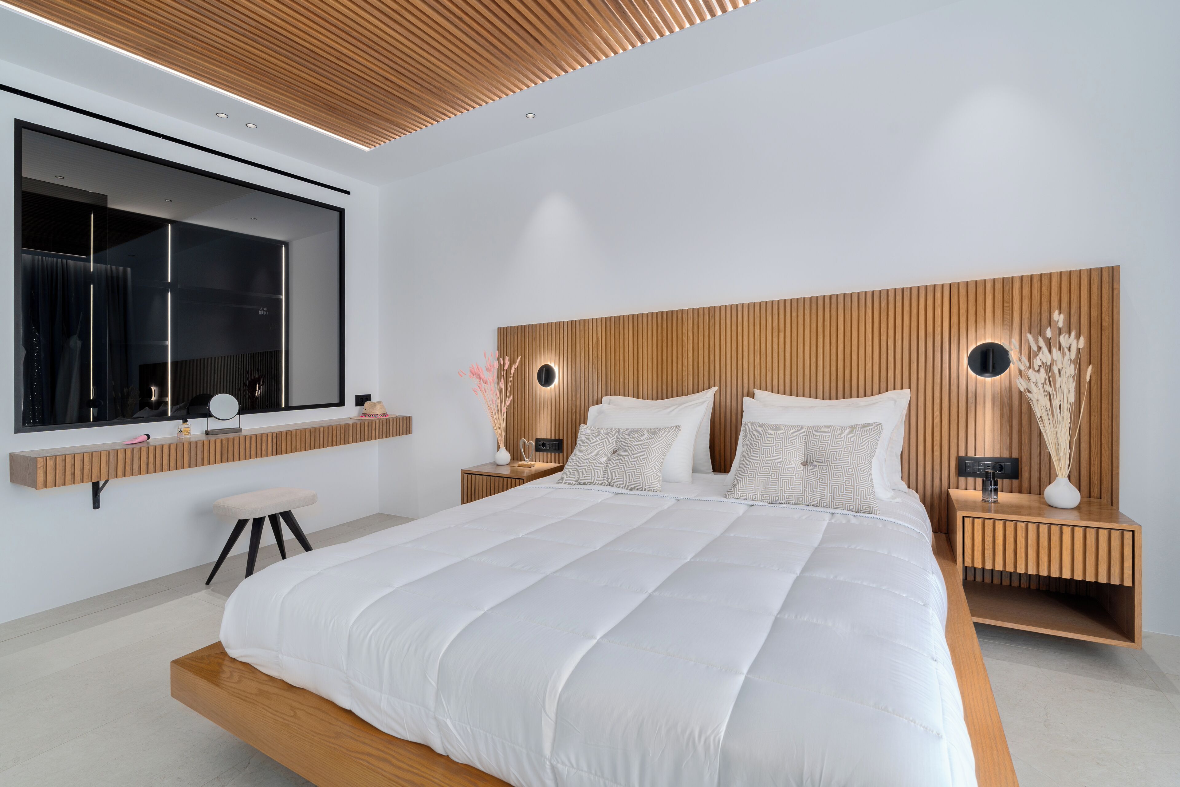 A modern bedroom with a large bed, wooden headboard, bedside tables with lamps, and a large mirror.