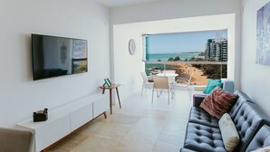 Una sala de estar moderna con televisión de pantalla plana, un sofá, un comedor y un balcón con vista a la playa y edificios.