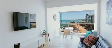 A modern living room with a flat-screen TV, a sofa, a dining area, and a balcony with a view of the beach and buildings.