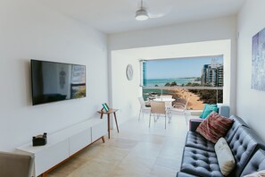 A modern living room with a flat-screen TV, a sofa, a dining area, and a balcony with a view of the beach and buildings.