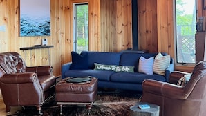 Interior acolhedor de uma cabana com paredes de madeira, um sofá azul, poltronas de couro e uma TV na parede.