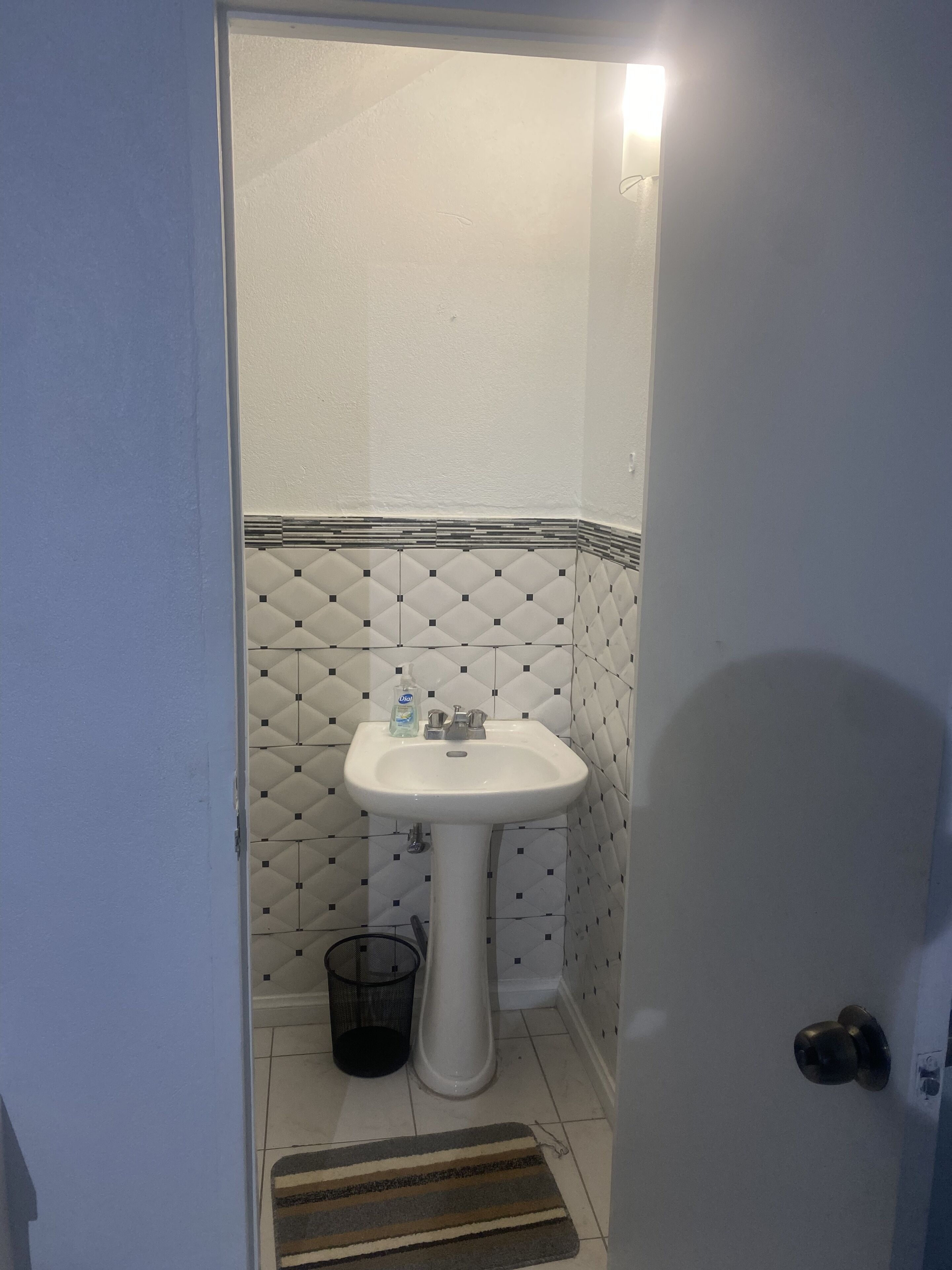 A bathroom with a white pedestal sink, a black trash bin, and a striped doormat.
