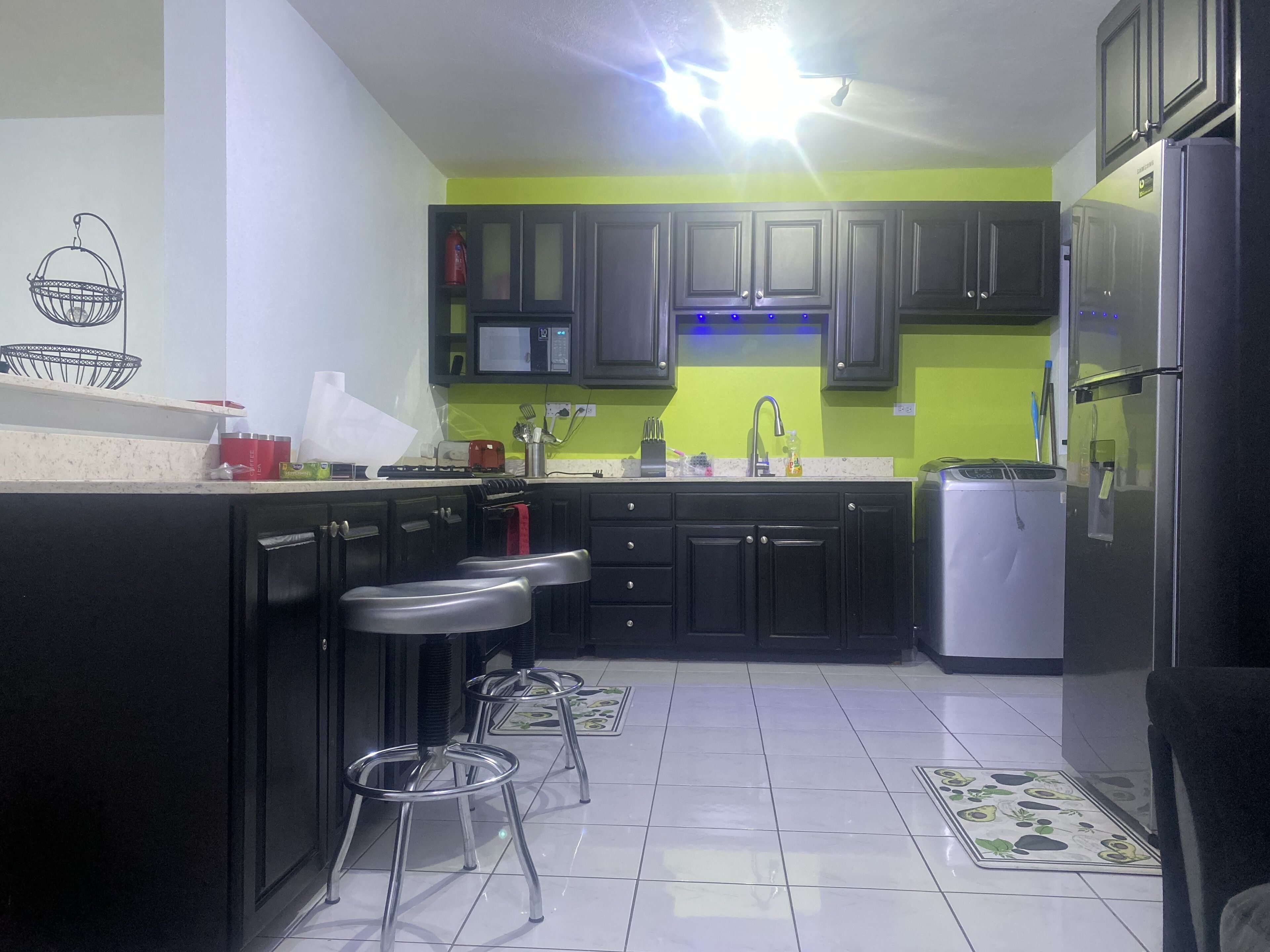 A kitchen with dark cabinets, a stainless steel refrigerator, a microwave, and a sink. There are two metal stools and a patterned rug on the floor.
